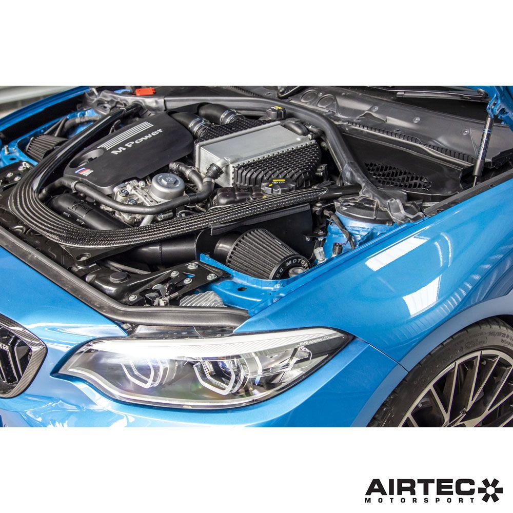 Airtec Motorsport induction kit installed in a BMW M2 Competition, M3, and M4 engine bay with air filters and intake tubing visible.
