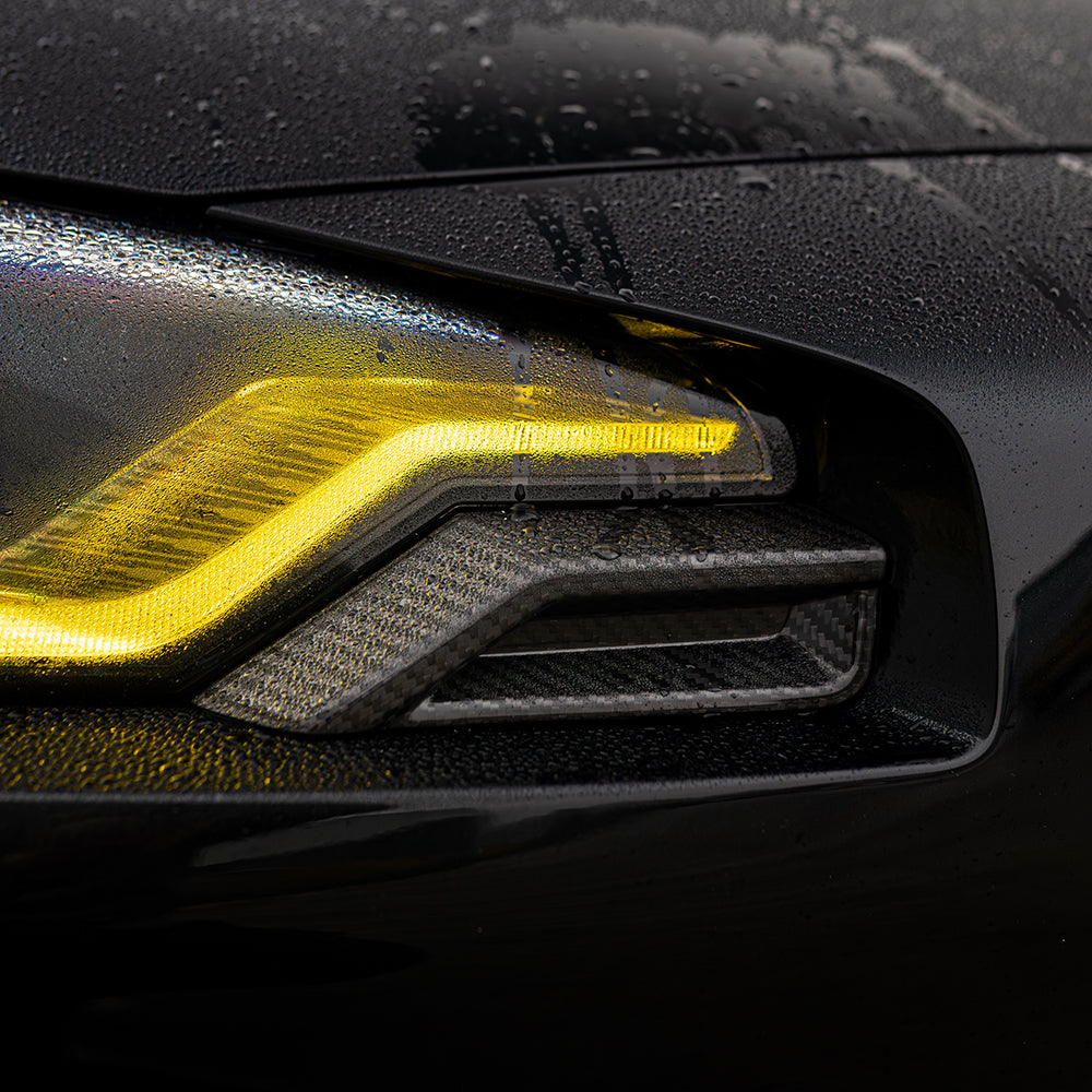 BMW G87 M2 carbon fiber front headlight add-on accessory by CT Carbon, shown in a close-up image highlighting the lightweight material.